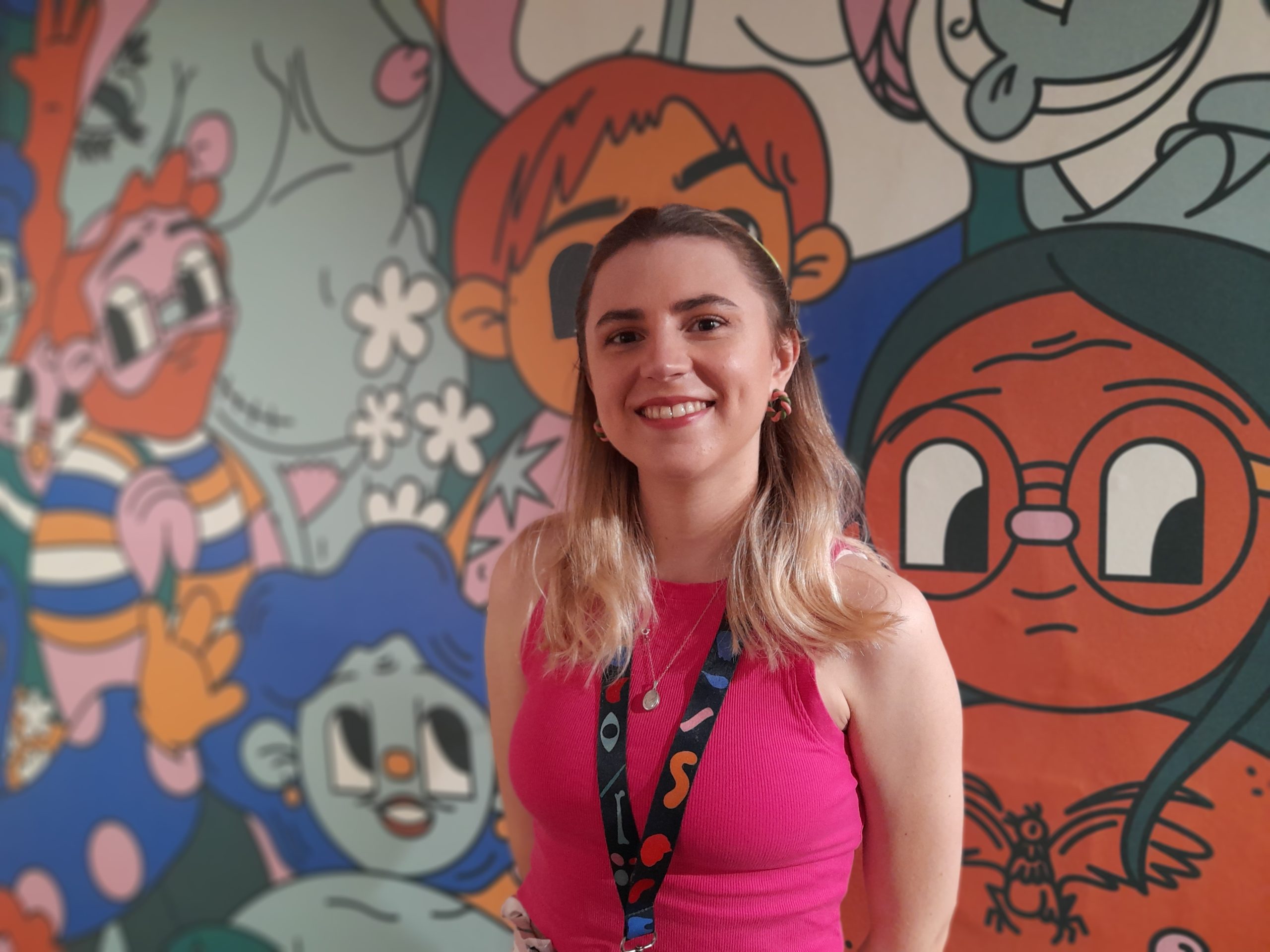 A young blonde woman in pink top against background of cartoon characters. 