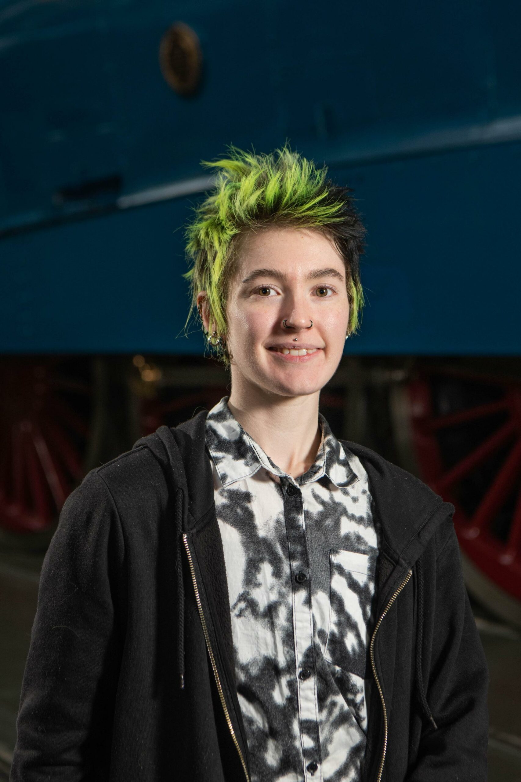 A young woman with short green hair. She is wearing a patterned shirt and black hoodie.