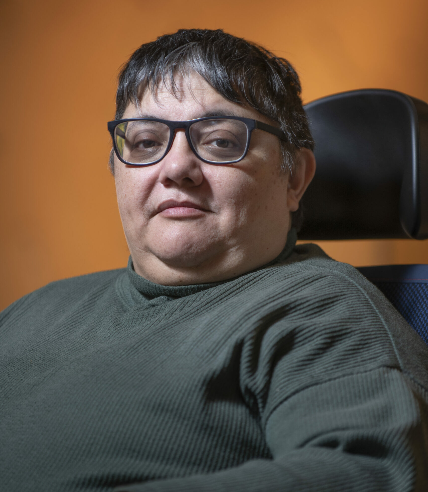 Suchi is a white woman with brown short hair who wears glasses. She is wearing a green jumper and sitting in a wheelchair.