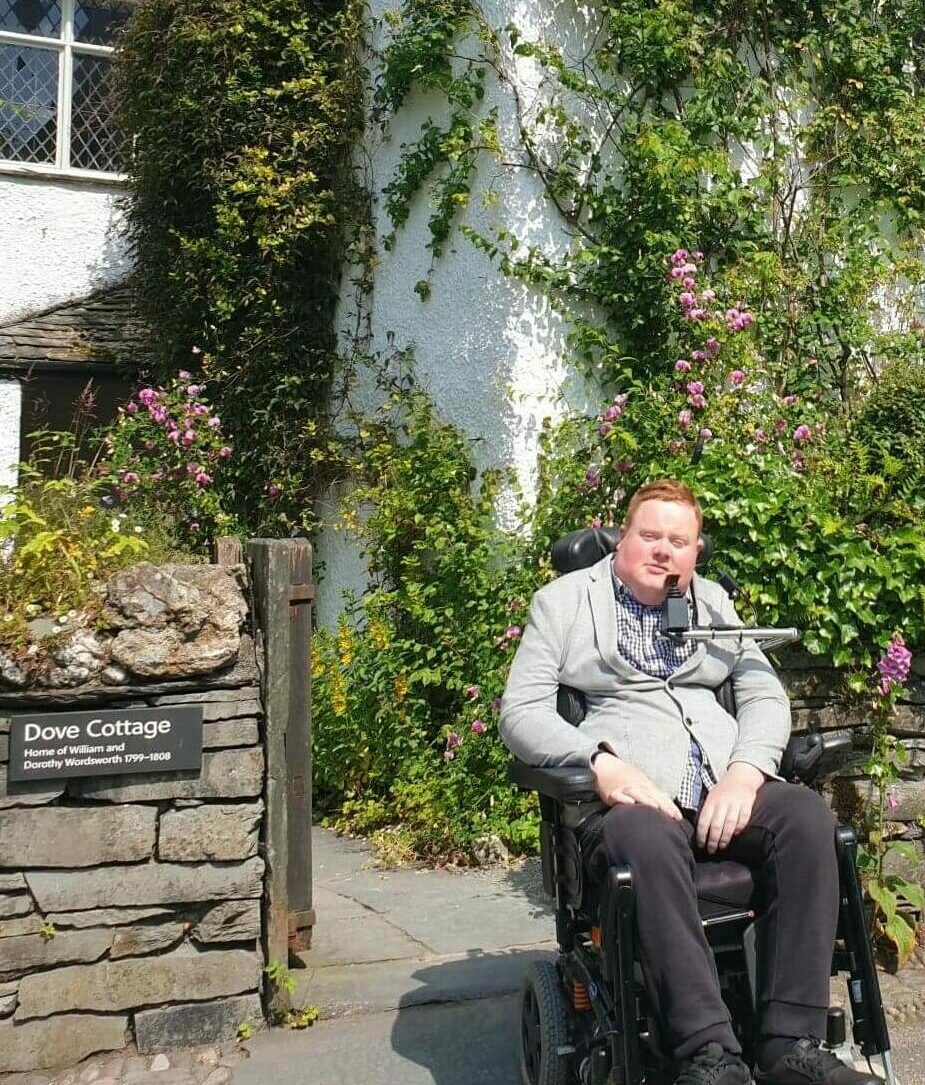Will Clark is a ginger man wearing a grey jumper and sitting in a wheelchair. He is sitting in front of Wordsworth's home, Dove Cottage.